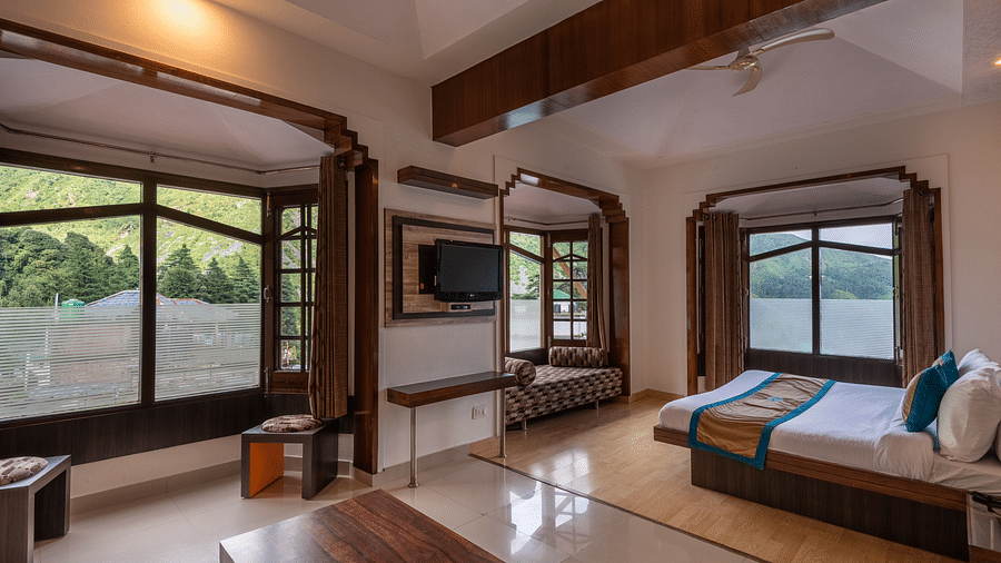 A suite with large windows offering a mountain view, a TV, a comfortable couch, and a king size bed with vibrant cushions - DLS Meghavan Resort, Dharamshala