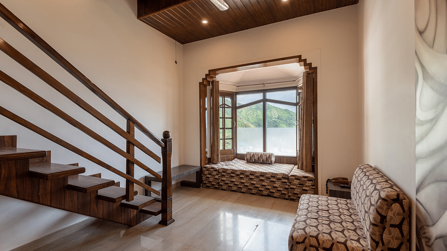 A room with wooden stairs, a seating area by a large window offering a mountain view, and a comfortable couch - DLS Meghavan Resort, Dharamshala