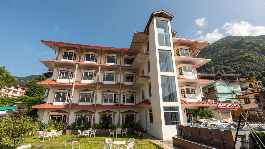 A resort in Bhagsu with a white and red facade, surrounded by outdoor seating in a garden area. The backdrop features green hills and nearby buildings - DLS Meghavan Resort, Dharamshala