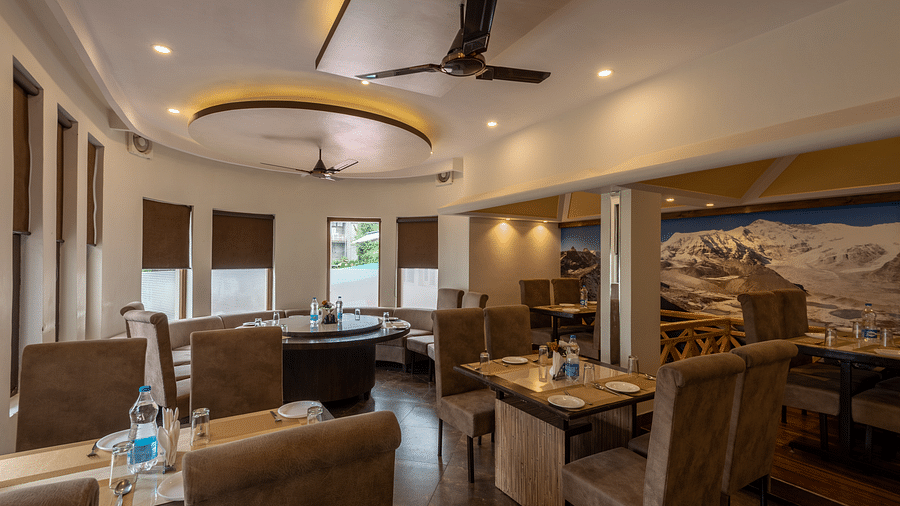 A restaurant with beige seating, tables set with plates, glasses, and cutlery. The space features modern lighting, ceiling fans, and a large mountain mural on the wall -  DLS Meghavan Resort, Dharamshala