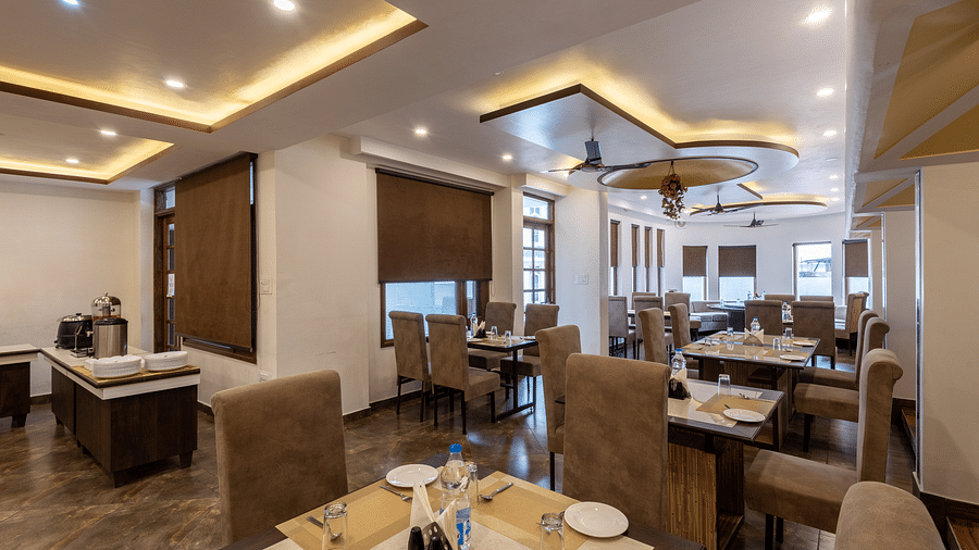 A restaurant with beige seating, tables set with plates, glasses, and cutlery. The space features modern lighting, brown window blinds, and a buffet area - DLS Meghavan Resort, Dharamshala