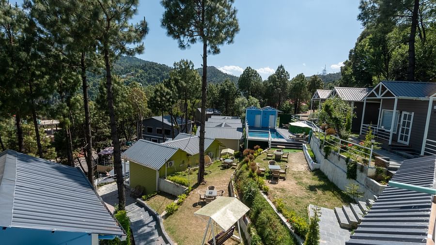 Daytime view of a hillside resort with charming cottages, manicured gardens, a swing set, and tall pine trees, set against a backdrop of forested mountains - DLS Nature Trinket Resort, Dalhousie.