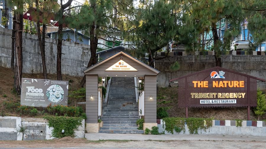 Entrance to a resort named 'The Nature Trinket Regency' featuring a staircase leading uphill through a small wooden-roofed gate, flanked by two large wooden signs and surrounded by tall trees and greenery.