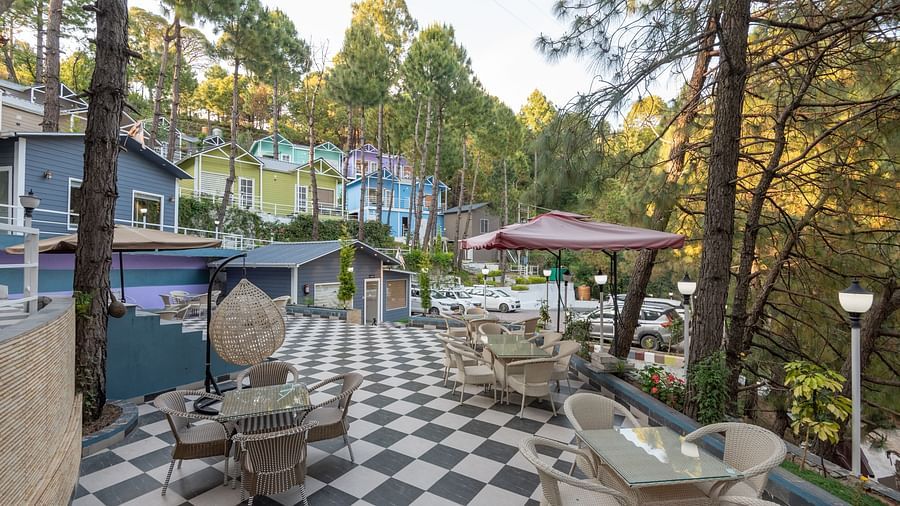 Elevated outdoor seating area with checkered black-and-white flooring, glass-top tables and wicker chairs, surrounded by tall pine trees and colourful hillside buildings under soft daylight - DLS Nature Trinket Resort, Dalhousie.
