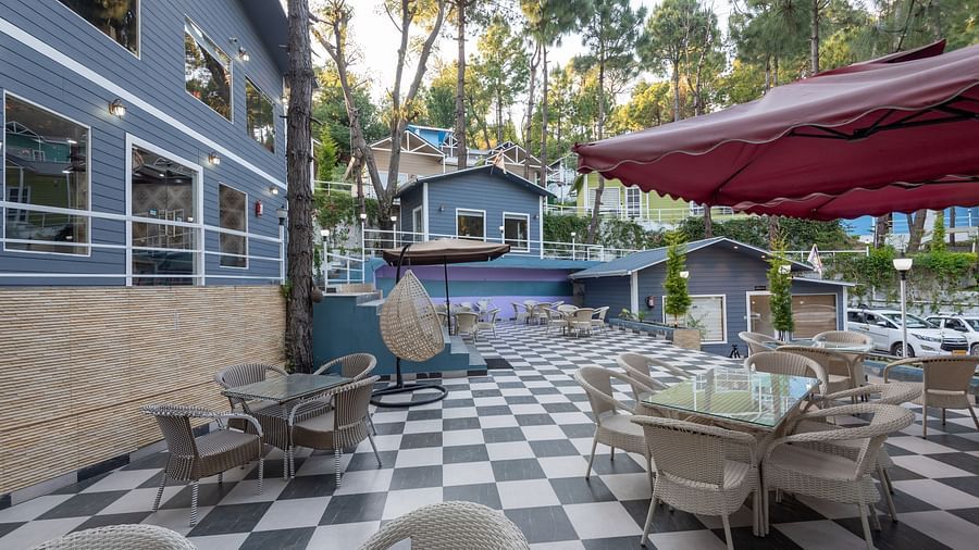 An outdoor seating area with wicker chairs and glass-top tables on a black-and-white checkered floor, shaded by large maroon umbrellas, surrounded by modern buildings and tall trees in the background - DLS Nature Trinket Resort, Dalhousie.