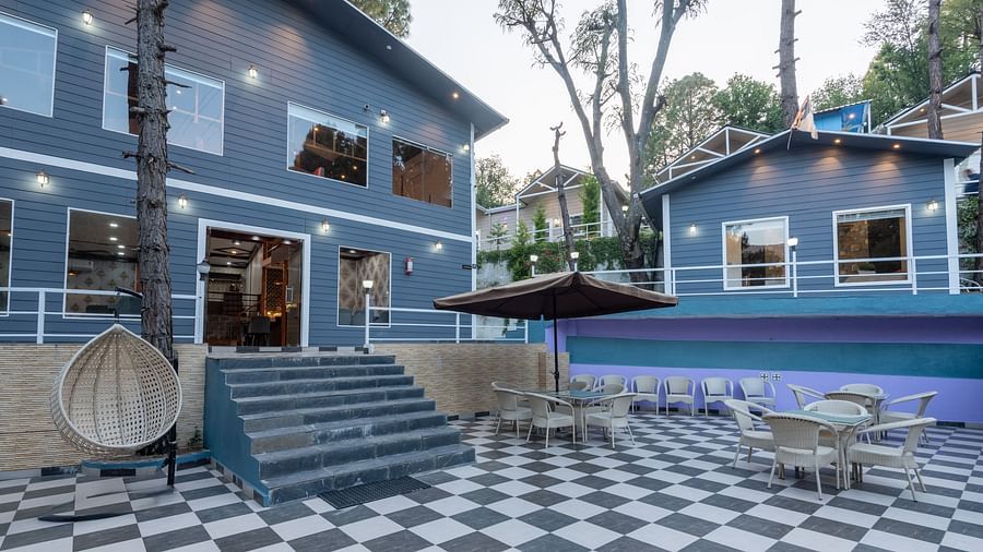 Outdoor patio area with black-and-white checkered flooring, modern blue buildings in the background, wicker chairs and tables, a hanging swing chair, and large umbrellas under tall pine trees - DLS Nature Trinket Resort, Dalhousie.