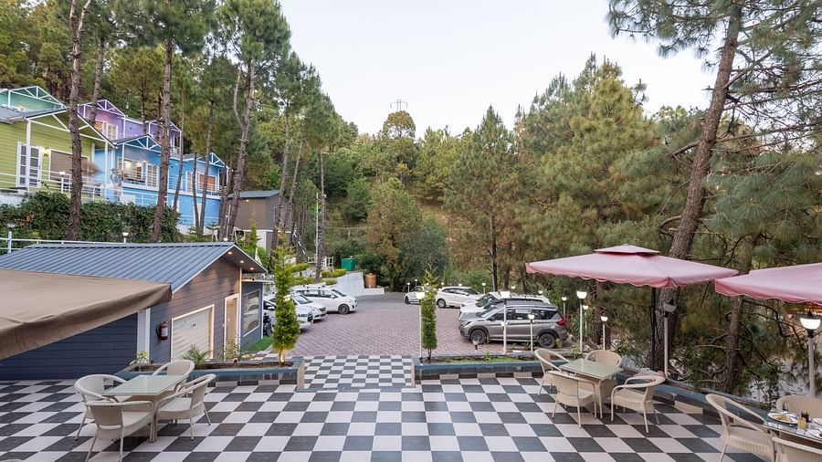 Outdoor restaurant seating area with black and white checkered flooring, surrounded by pine trees, parked cars, and colourful buildings in the background - DLS Nature Trinket Resort, Dalhousie.