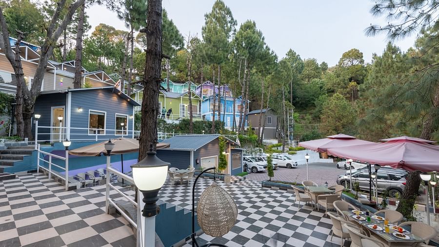 Outdoor dining area with black and white checkered flooring, beige tables with red umbrellas, surrounded by pine trees and colourful hillside buildings, creating a cosy, forested ambience - DLS Nature Trinket Resort, Dalhousie.