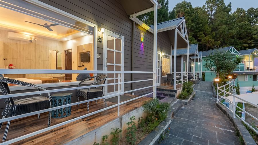 Evening view of a stone pathway leading past modern wooden cottages with large windows, warmly lit interiors, and white railings, surrounded by greenery - DLS Nature Trinket Resort, Dalhousie.