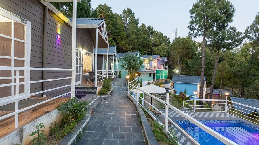 Scenic view of a hillside resort with cottage-style accommodations, a stone-paved walkway, and a swimming pool, surrounded by pine trees - DLS Nature Trinket Resort, Dalhousie.
