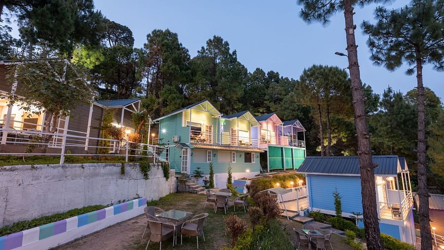  Twilight view of a hillside resort with colourful cottages, ambient lighting, garden seating areas, and tall pine trees nestled against a forested slope - DLS Nature Trinket Resort, Dalhousie.