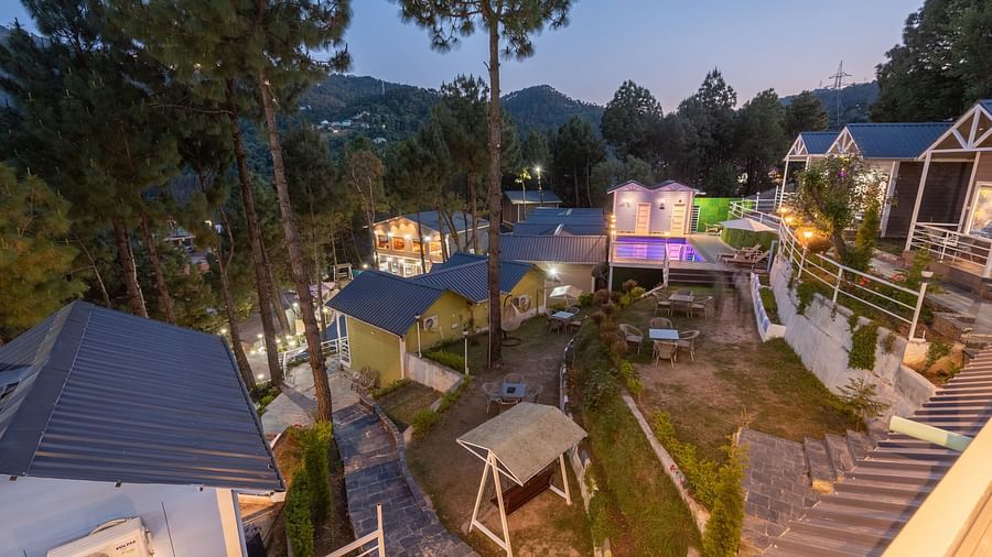  Elevated evening view of a hillside resort featuring cosy cottages, a central garden area with a swing, a lit-up swimming pool, and tall pine trees with mountain views in the background - DLS Nature Trinket Resort, Dalhousie.