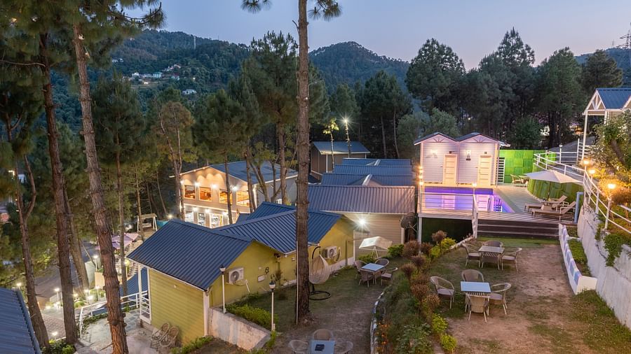 Elevated twilight view of a serene hillside resort with cosy cottages, an illuminated swimming pool, outdoor seating, and tall pine trees set against a backdrop of forested hill - DLS Nature Trinket Resort, Dalhousie.