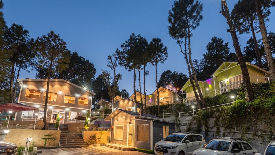 Twilight view of a hillside resort with warmly lit buildings, parked cars, and tall pine trees silhouetted against the evening sky - DLS Nature Trinket Resort, Dalhousie.