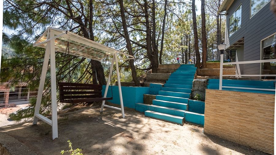 Outdoor patio with a wooden swing, turquoise steps, and a modern house surrounded by tall trees - DLS Nature Trinket Resort, Dalhousie.