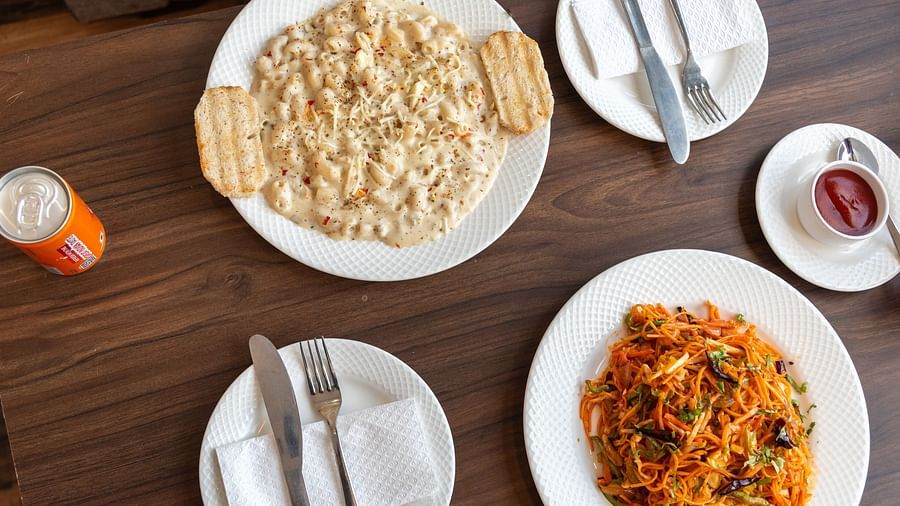 A delicious spread featuring a plate of creamy pasta with crispy breadsticks, alongside a plate of colourful noodles, and a serving of ketchup, complemented by a refreshing orange drink - DLS Nature Trinket Resort, Dalhousie.