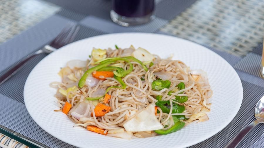 A white plate filled with stir-fried noodles and mixed vegetables such as bell peppers, carrots, and cabbage, placed on a grey placemat with utensils beside it - DLS Nature Trinket Resort, Dalhousie.