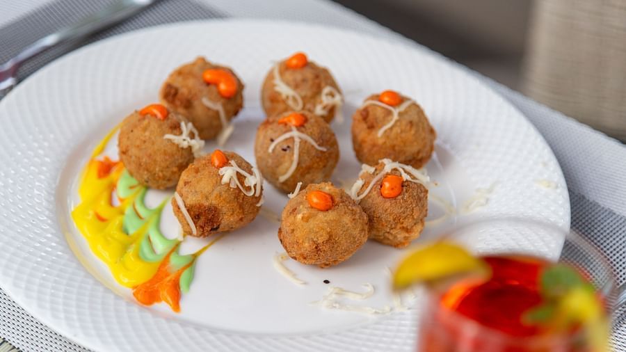 A plate of round snack balls garnished with orange toppings and grated cheese, surrounded by a colourful drizzle of sauce. A refreshing drink is partially visible in the background - DLS Nature Trinket Resort, Dalhousie.