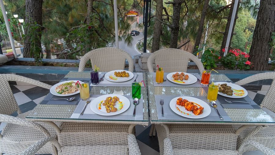 Outdoor dining setup with plates of food and drinks on a glass table, surrounded by wicker chairs and trees in the background - DLS Nature Trinket Resort, Dalhousie. 
