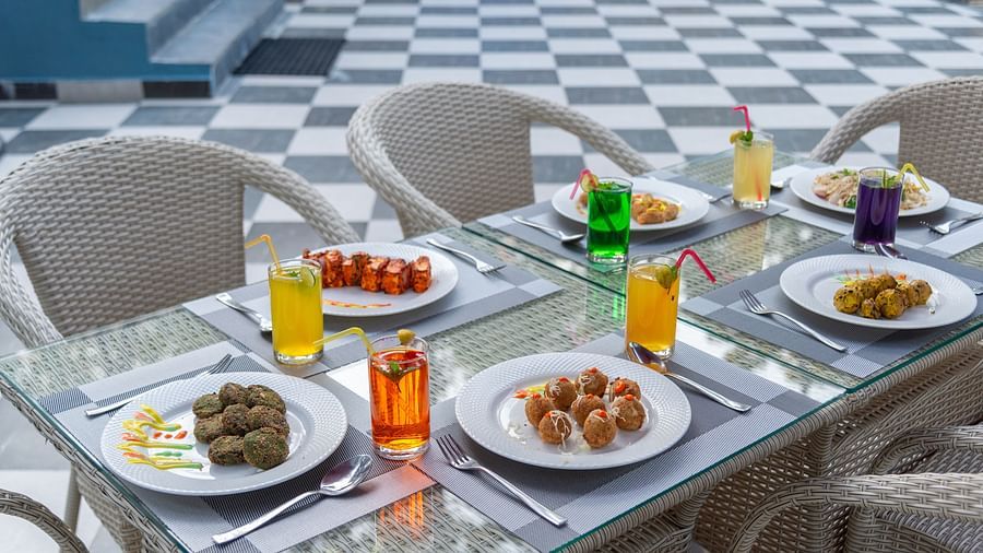 A stylishly set outdoor dining table with plates of appetising snacks and starters, accompanied by colourful drinks in a variety of hues, surrounded by a checkered floor and fresh ambience - DLS Nature Trinket Resort, Dalhousie.