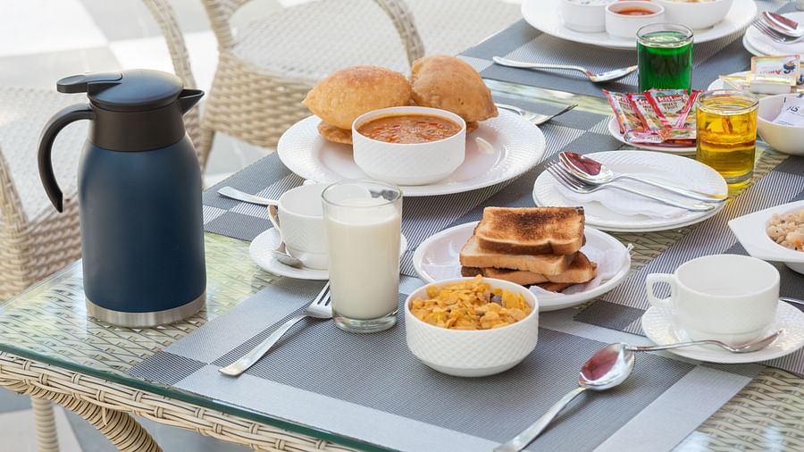Close-up of a breakfast table set with cereal, toast, buns, tea or coffee, juice, and condiments on a glass-top table with wicker chairs - DLS Nature Trinket Resort, Dalhousie.