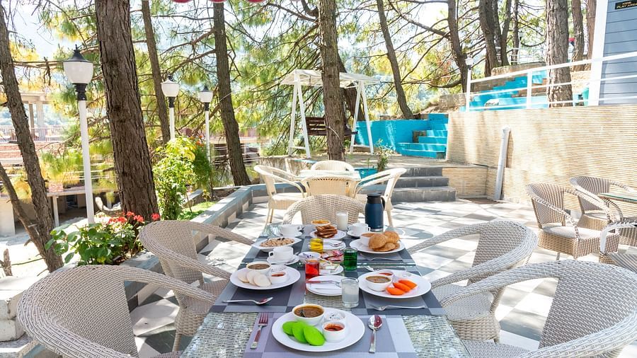 Spacious outdoor dining area set for breakfast, featuring a glass-top table with assorted dishes, surrounded by white chairs under a red canopy, nestled among tall trees and natural greenery - DLS Nature Trinket Resort, Dalhousie.