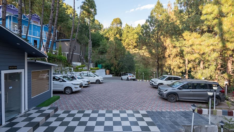 View of a paved parking lot with multiple cars parked, bordered by a forest of tall pine trees and colourful buildings, with a black-and-white checkered tile area in the foreground - DLS Nature Trinket Resort, Dalhousie.
