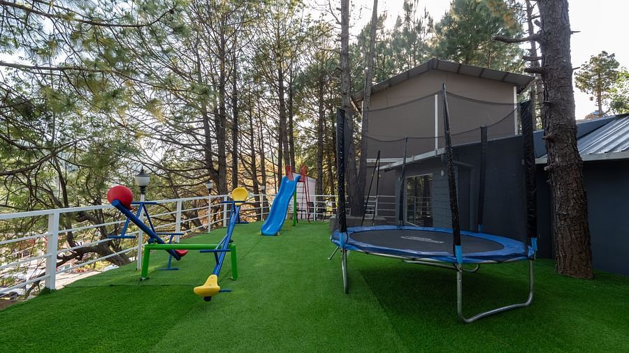 A children's play area on green artificial turf with a blue trampoline, a blue slide, and a colourful seesaw, surrounded by tall pine trees and a safety railing, next to a wooden structure - DLS Nature Trinket Resort, Dalhousie.