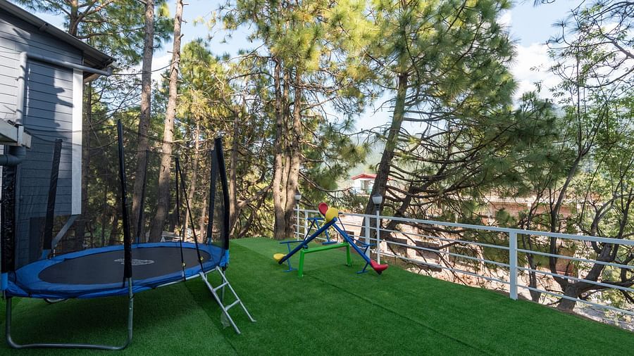 Outdoor children's play area with a trampoline, slide, and seesaw on artificial green turf, surrounded by trees and a white safety railing, located next to a grey building - DLS Nature Trinket Resort, Dalhousie.