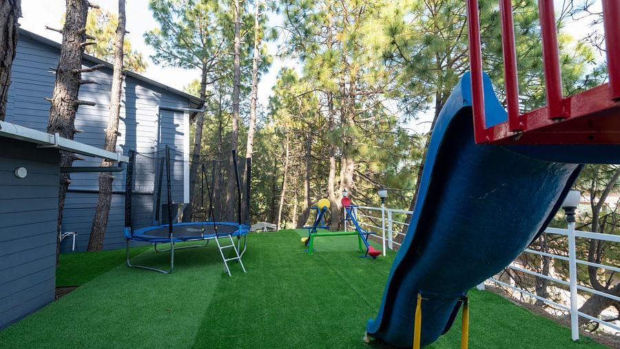  Outdoor children's play area with artificial grass, featuring a blue slide, trampoline, and colourful climbing equipment, surrounded by pine trees and adjacent buildings - DLS Nature Trinket Resort, Dalhousie.