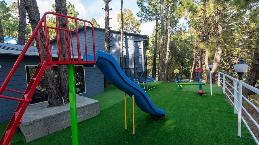 Outdoor playground with artificial grass, featuring a blue slide with red and green support structure, surrounded by pine trees and enclosed by a white railing - DLS Nature Trinket Resort, Dalhousie.