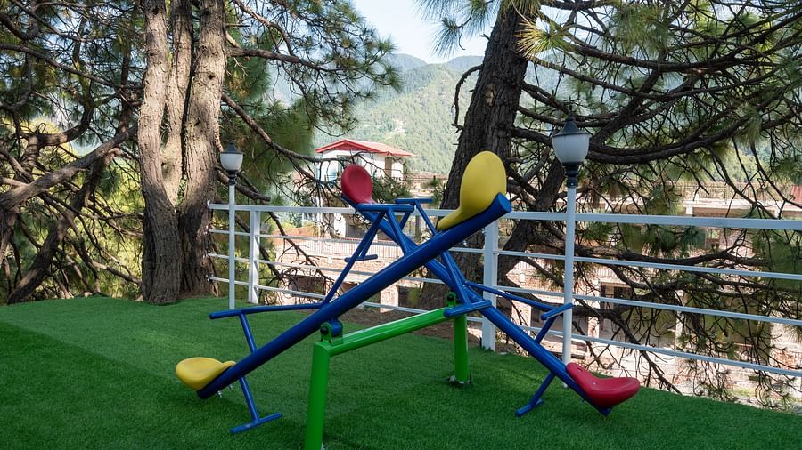  Colourful seesaw play equipment with red, yellow, and blue seats placed on artificial green turf, set against a backdrop of pine trees and mountain scenery - DLS Nature Trinket Resort, Dalhousie.