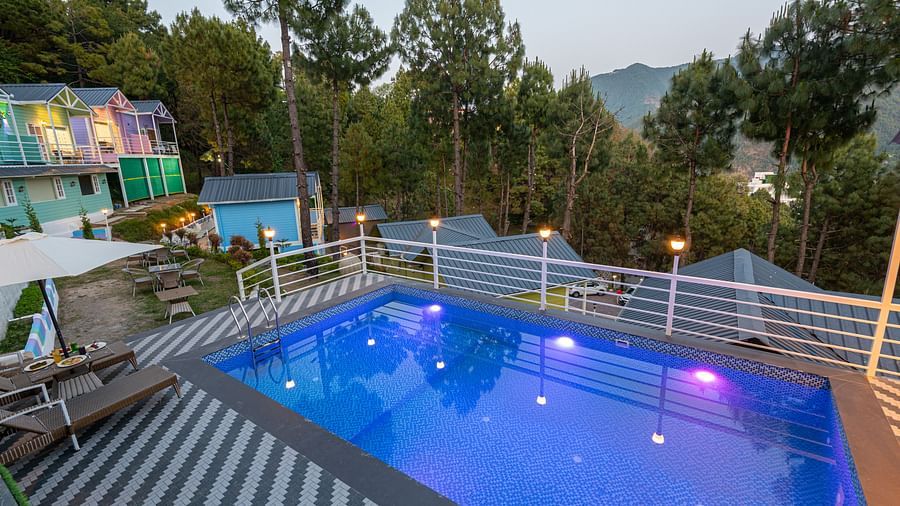 Elevated evening view of a swimming pool with colourful underwater lighting, surrounded by striped decking, lounge chairs, and vibrant hillside cottages amid pine trees and mountain scenery - DLS Nature Trinket Resort, Dalhousie.