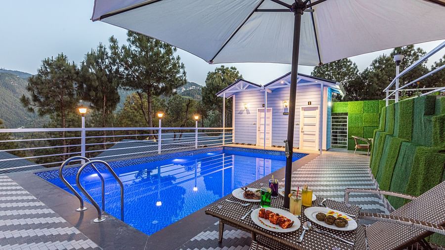 Evening poolside scene with a lit swimming pool, two lounge chairs under a large umbrella, and plates of food and drinks arranged on a table, set against a backdrop of trees and mountains - DLS Nature Trinket Resort, Dalhousie.