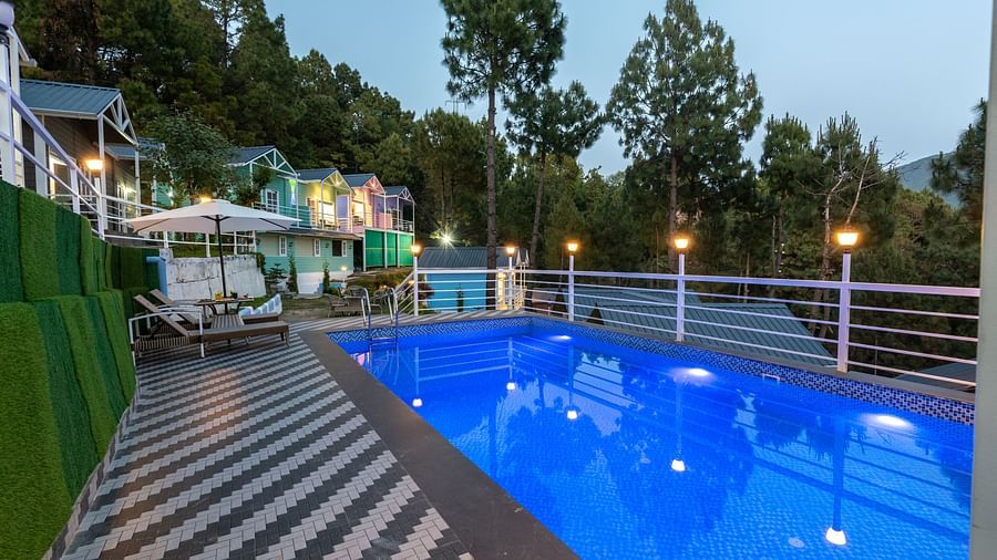 Illuminated swimming pool with glowing blue water in the evening, surrounded by patterned tile flooring, lounge chairs, and colourful cottages nestled among pine trees - DLS Nature Trinket Resort, Dalhousie.