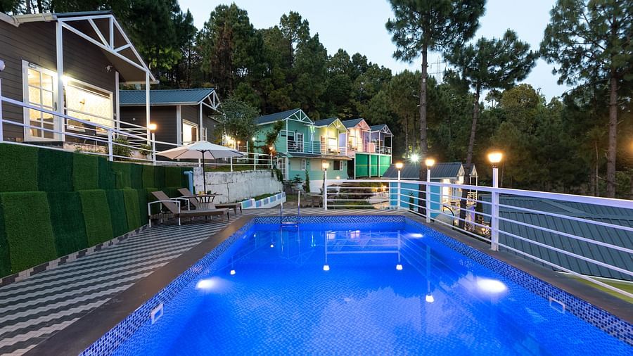 Tranquil evening view of a lit swimming pool surrounded by safety railings and striped flooring, with cosy hillside cottages and tall pine trees in the background - DLS Nature Trinket Resort, Dalhousie.