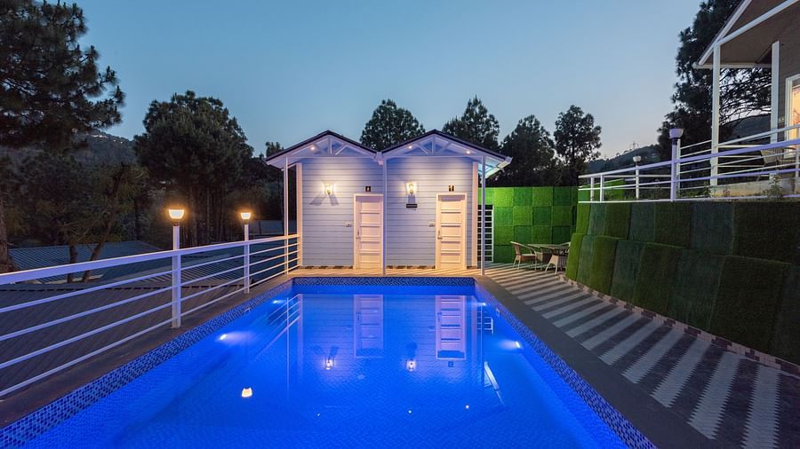  Evening view of a glowing blue swimming pool with modern lighting, flanked by white railings and striped tile flooring, with two illuminated poolside cabins in the background and surrounded by trees - DLS Nature Trinket Resort, Dalhousie. 