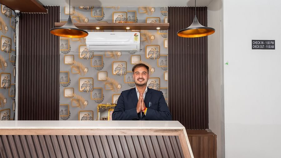  A smiling man in a dark suit stands behind a modern reception desk with folded hands in a greeting gesture, set against a backdrop of patterned wallpaper, pendant lights, and a wall-mounted air conditioner - DLS Nature Trinket Resort, Dalhousie.