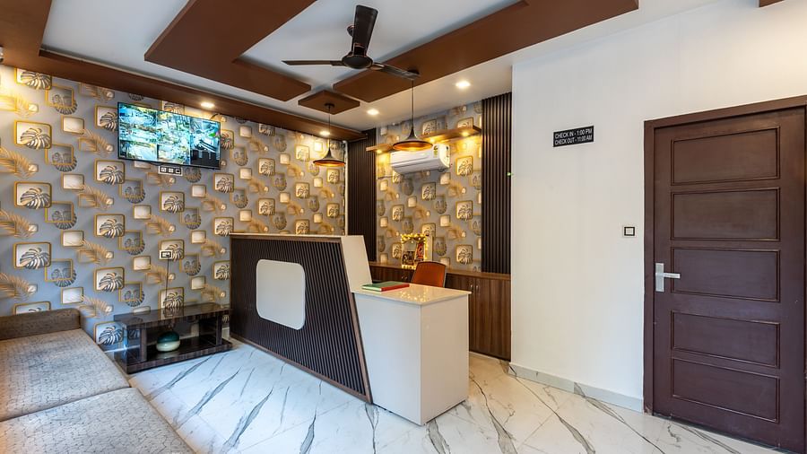 A stylish reception room with a white marble floor, wood-accented ceiling panels with a ceiling fan, patterned wallpaper, a small front desk, wall-mounted TV, and a dark brown door on the right - DLS Nature Trinket Resort, Dalhousie.