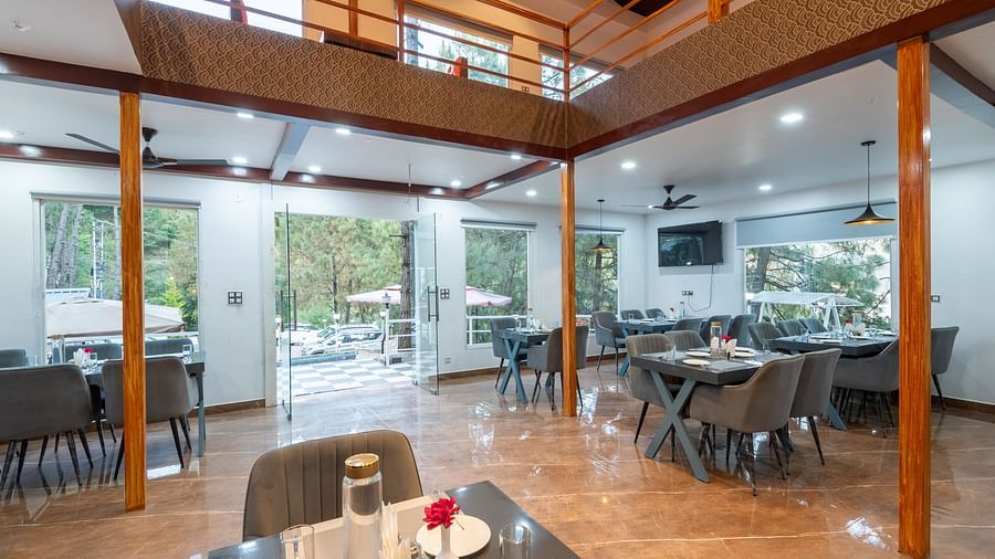 Bright and modern restaurant dining area with neatly arranged tables, grey chairs, large windows, and an open mezzanine level - DLS Nature Trinket Resort, Dalhousie.