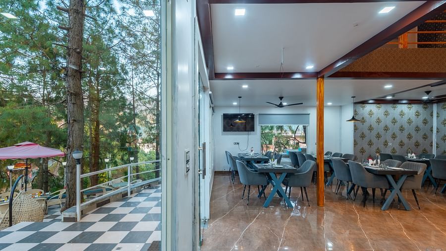 The interior of a restaurant is shown with tables and chairs. Through the open doorway, there's a view of a hillside - DLS Nature Trinket Resort, Dalhousie.