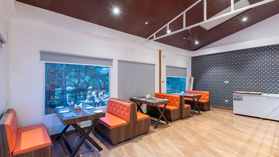 A brightly lit restaurant interior features wooden floors, a dark brown ceiling, tables with place settings, orange cushioned booths and chairs, and large windows with grey blinds - DLS Nature Trinket Resort, Dalhousie.