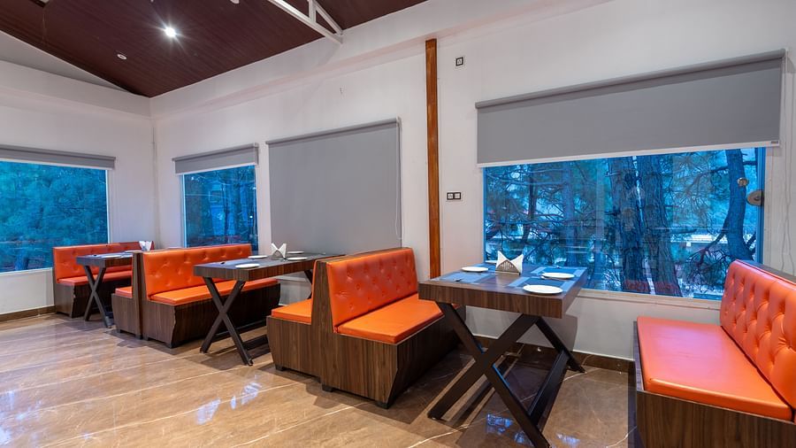 A brightly lit restaurant interior features wooden floors, a dark brown ceiling, tables with place settings, and orange cushioned booths and chairs situated near large windows with grey blinds - DLS Nature Trinket Resort, Dalhousie.