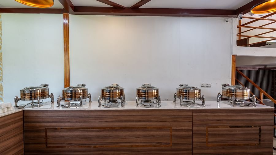  Buffet setup with stainless steel chafing dishes arranged on a wooden counter against a plain wall in a modern restaurant - DLS Nature Trinket Resort, Dalhousie.
