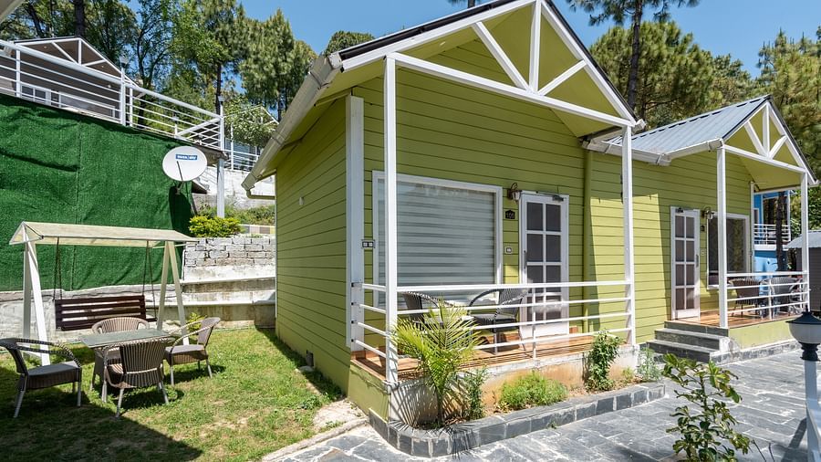 Bright green wooden cottages with white trim, set along a stone pathway, featuring a grassy outdoor seating area with a swing and surrounded by tall trees - DLS Nature Trinket Resort, Dalhousie.