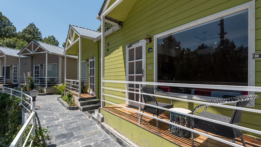  Row of modern green-painted cottages with white railings, private porches, outdoor seating, and a stone-paved walkway surrounded by trees - DLS Nature Trinket Resort, Dalhousie.