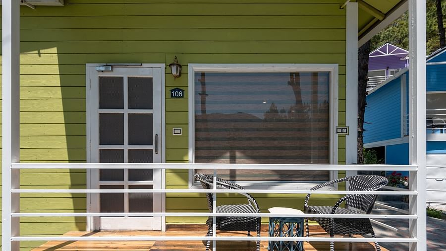  Front porch of a green wooden cottage with white railing, featuring a small outdoor seating area with a table and chairs, and a door and large window behind - DLS Nature Trinket Resort, Dalhousie.
