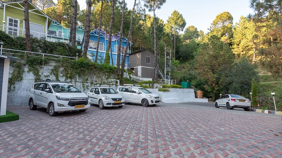  Spacious parking area with several white cars parked on a tiled surface, set against a backdrop of colourful hillside cottages and tall pine trees - DLS Nature Trinket Resort, Dalhousie.