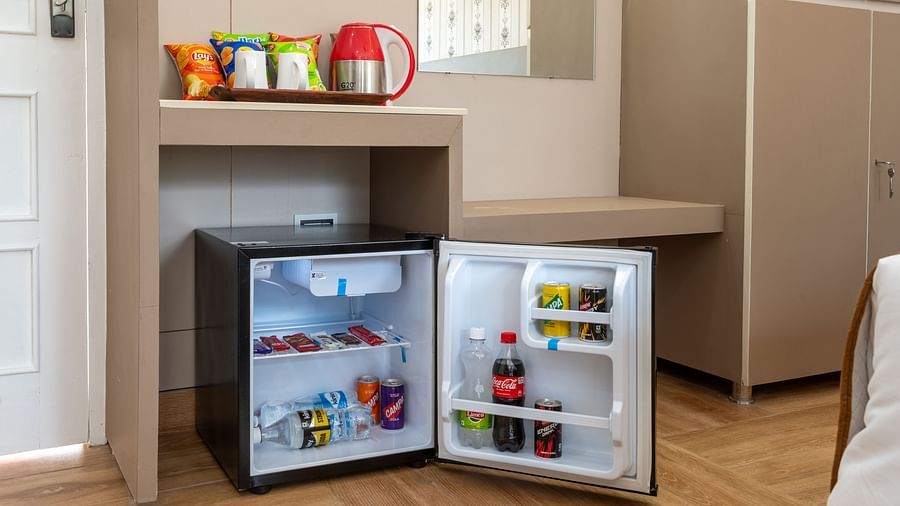  Small open minibar fridge in a hotel room, stocked with beverages and snacks, with a red electric kettle, cups, and complimentary items on the counter above - DLS Nature Trinket Resort, Dalhousie.