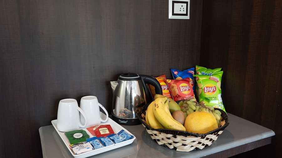 Hospitality tray on a counter featuring a kettle, two white cups, a selection of tea and coffee sachets, and a fruit basket with bananas, mangoes, and crisps - DLS Nature Trinket Resort, Dalhousie.